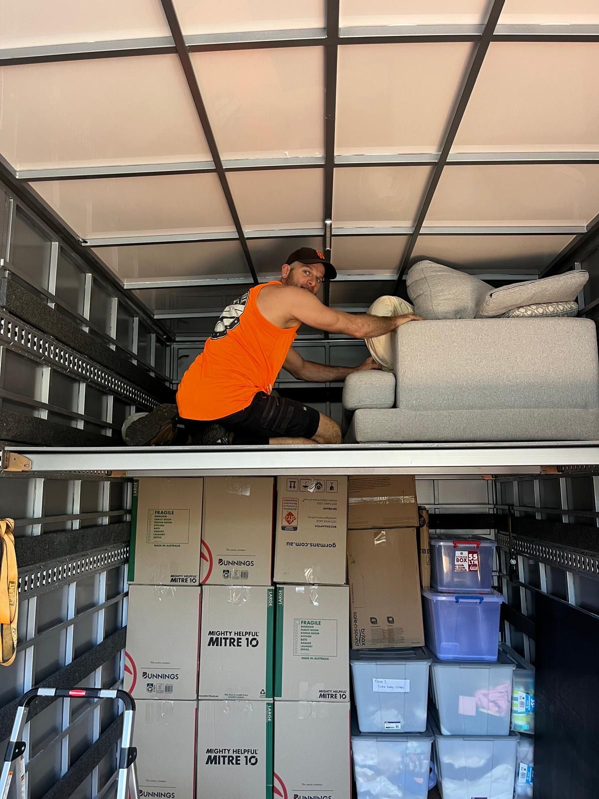 Man in orange shirt loading furniture into a moving truck, boxes stacked below— Ready To Move in Mullumbimby, NSW