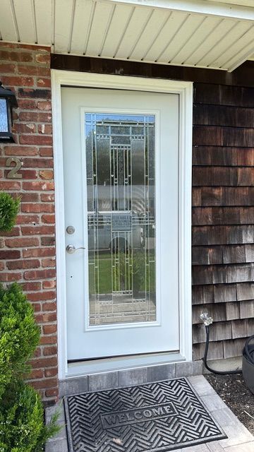 White front door with glass panel and metal security grate, set beside brick and shingle siding with a doormat.