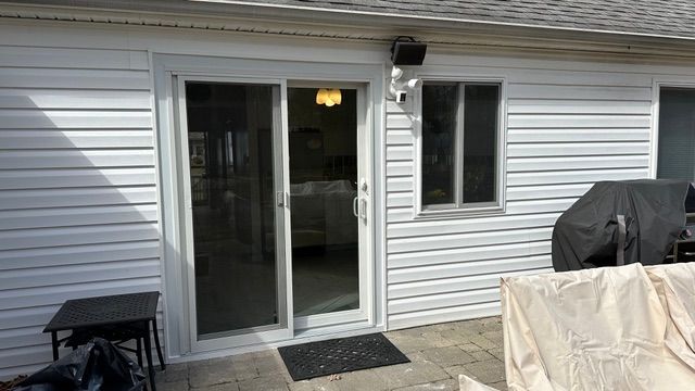 Back patio with sliding glass door, small table, grill covered with gray tarp, and a covered chair.