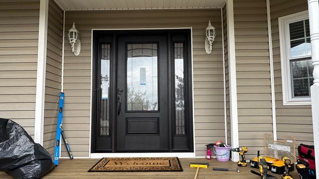 Front porch with black front door, tan siding, doormat, and cleaning supplies on the right