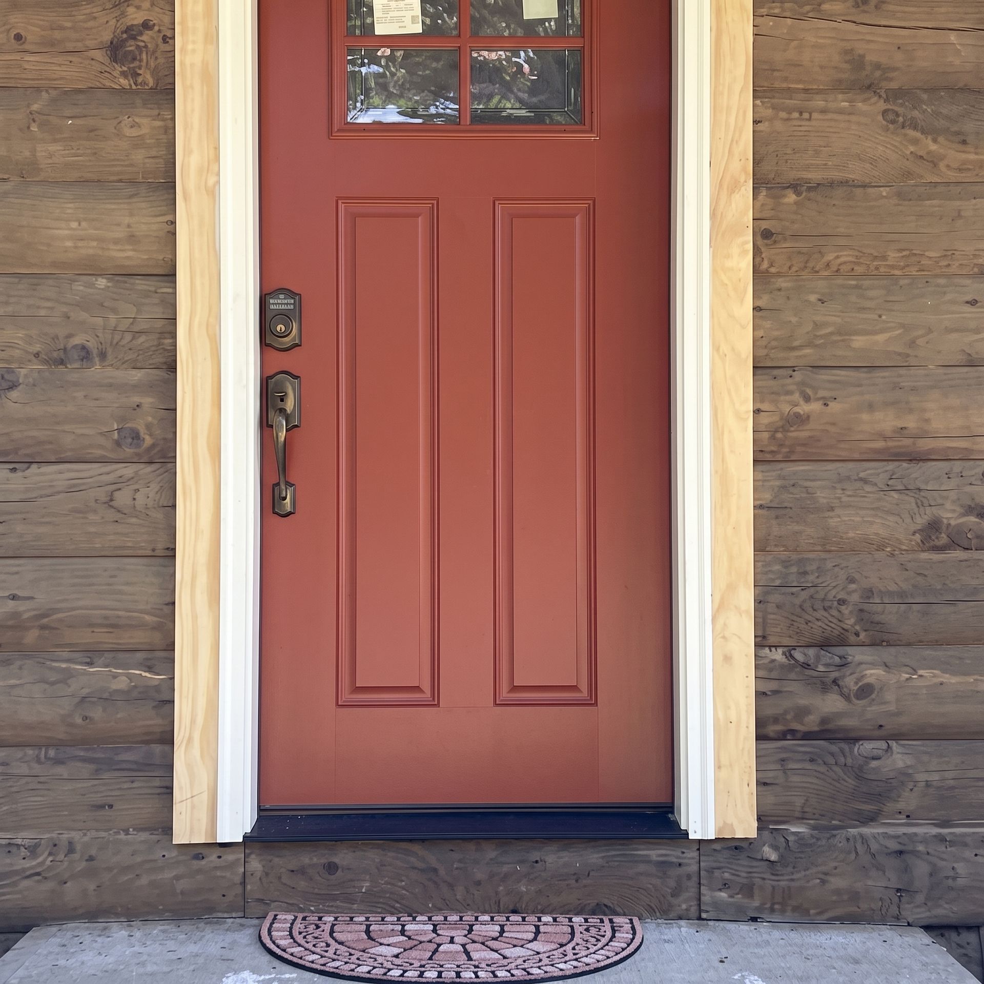 Red front door with a window panel, framed by light trim on a wood-sided house, with a welcome mat below.