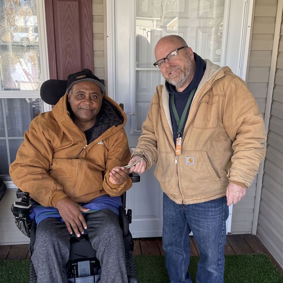 Two people standing on a porch, smiling and holding hands, one in a wheelchair.