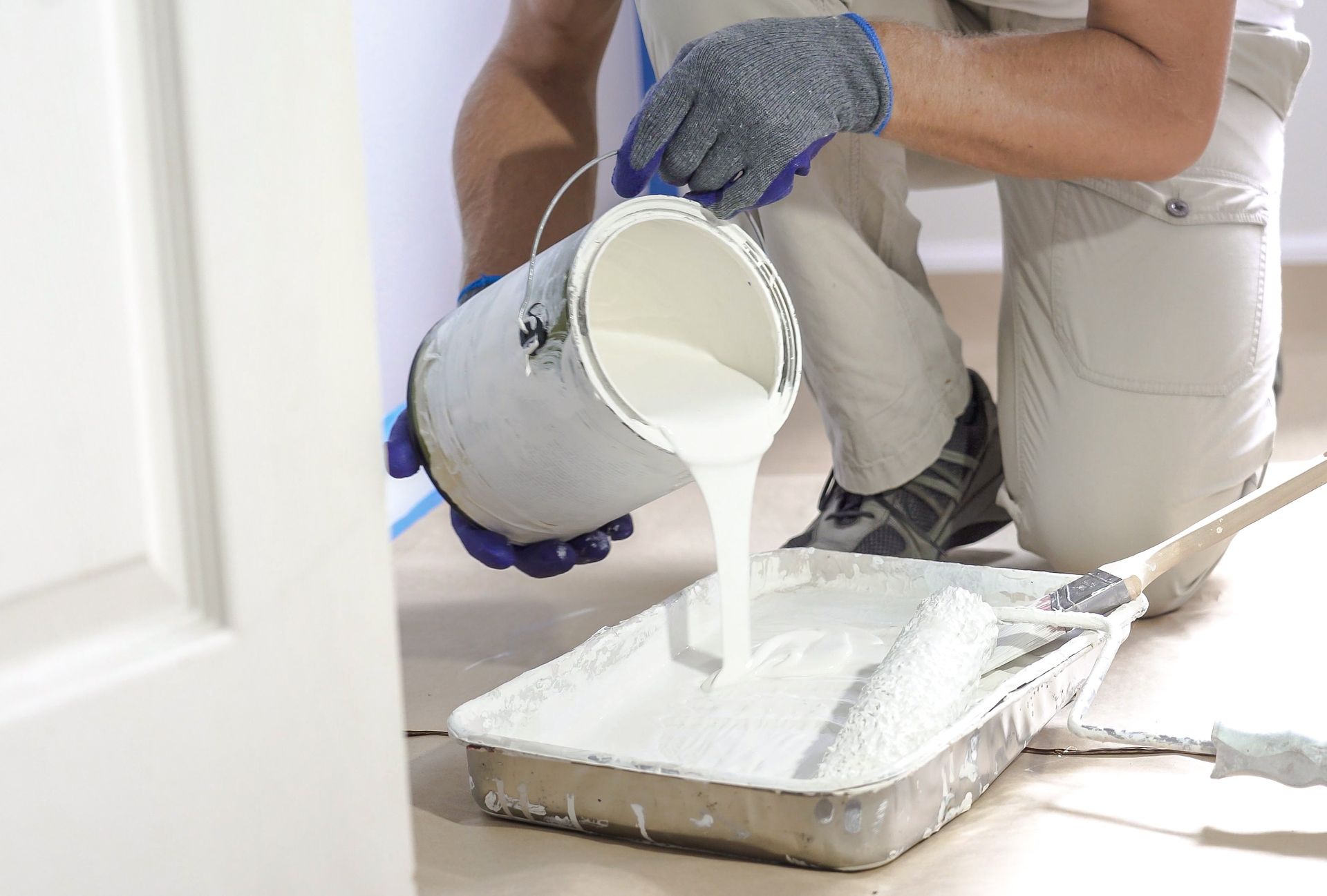 Person pouring white paint into a tray while kneeling on a floor indoors