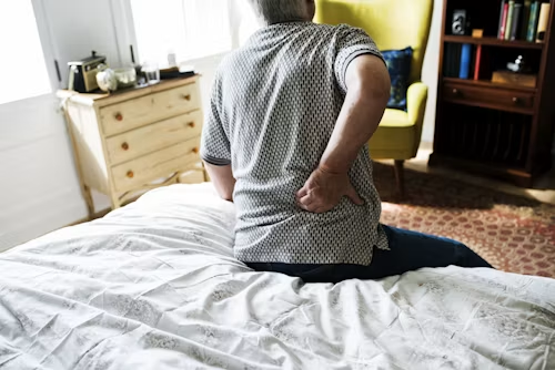 Person sitting on the edge of a bed in a bright bedroom, holding their lower back.