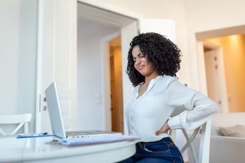 Woman sitting at a laptop, holding her lower back in pain, indoors.