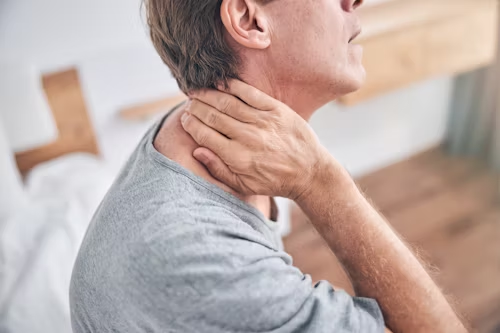 Man holding neck, appearing to be in pain. Indoors, wearing gray shirt.
