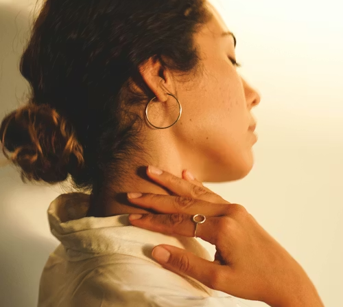 A person in profile wearing a thin gold hoop earring and a delicate gold ring, with their hand resting on their neck.