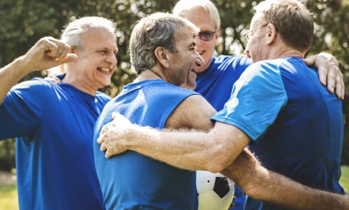 Four men in blue shirts huddle together, smiling, with a soccer ball. Outdoors.