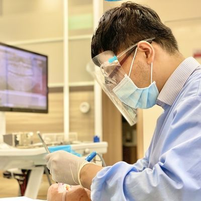 A dentist wearing a mask and a face shield is working on a patient 's teeth.