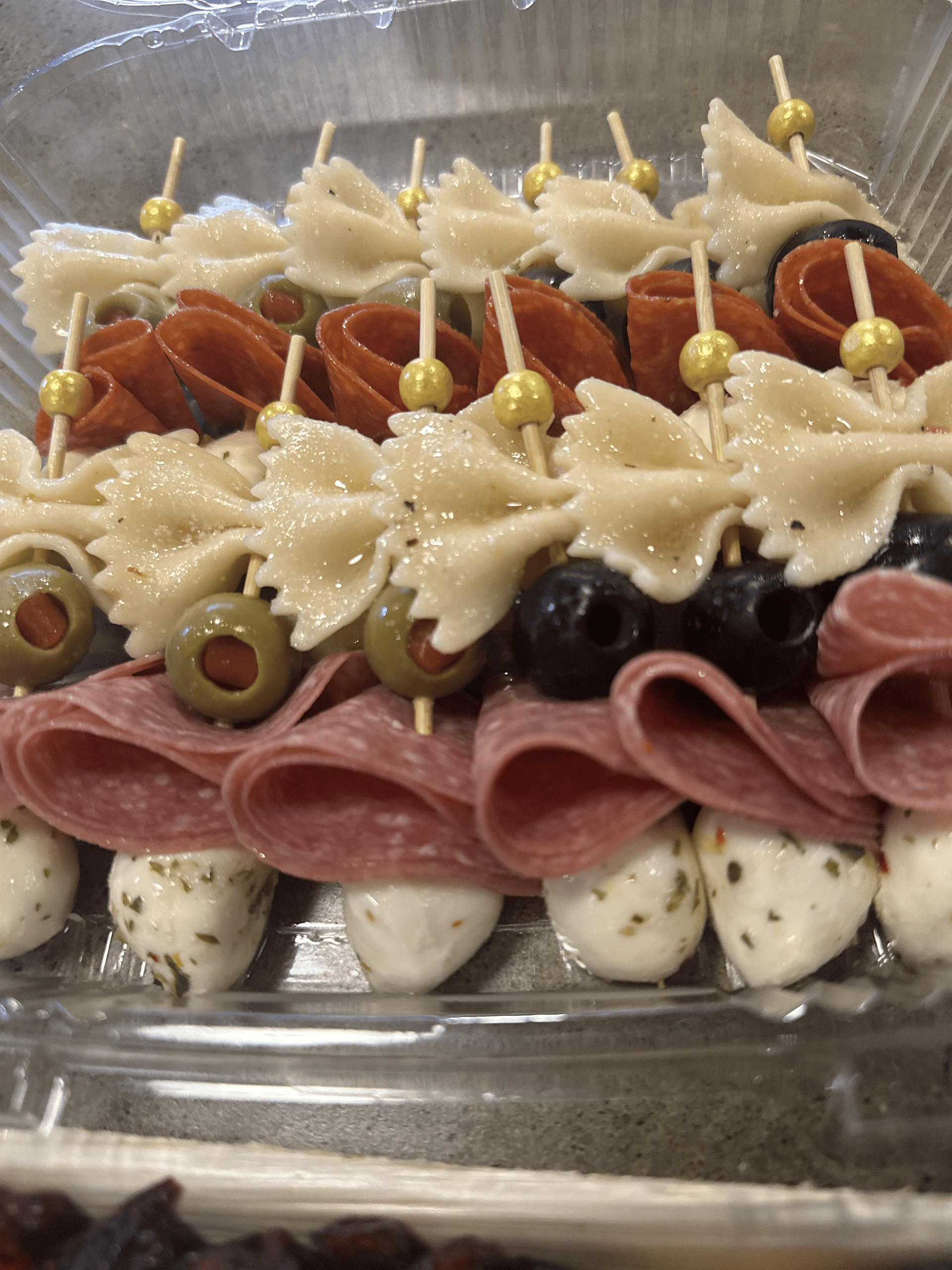 Appetizer skewers with pasta, olives, salami, and mozzarella balls in a clear container.