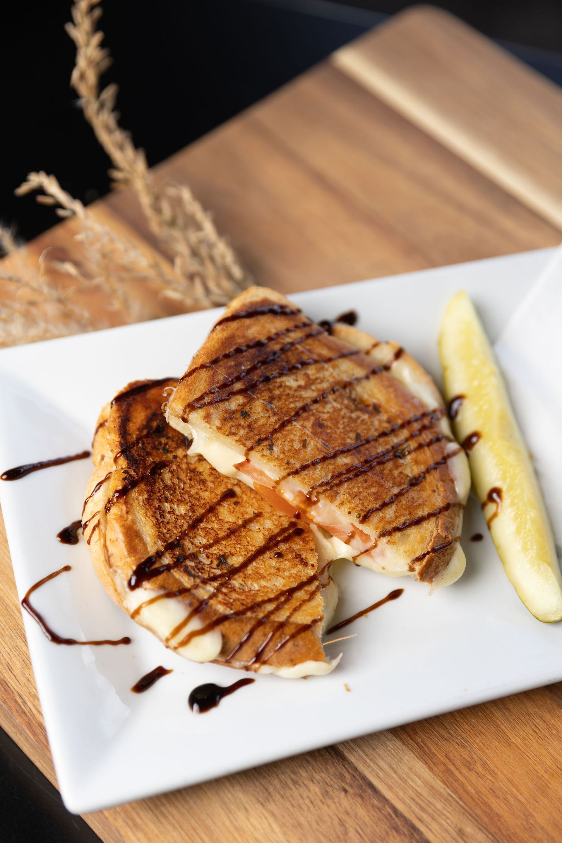 Grilled cheese sandwich drizzled with balsamic glaze, served with a pickle spear on a white plate.
