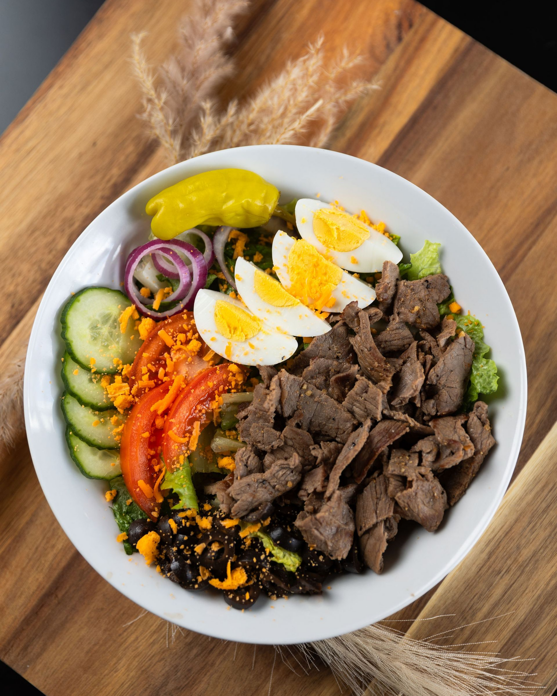 Salad bowl with sliced meat, eggs, vegetables, and a pepper on a wooden board.