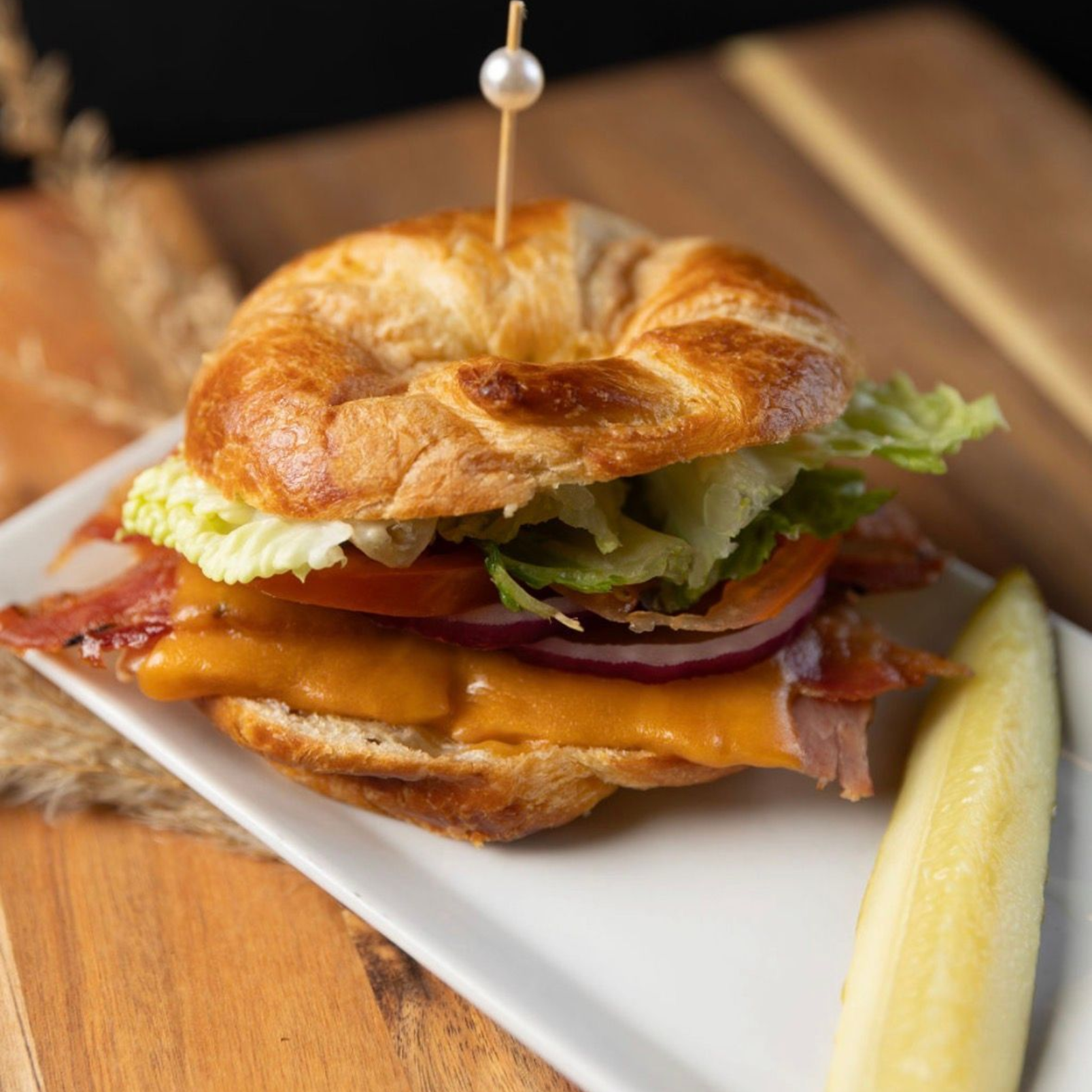 Croissant sandwich with cheese, bacon, tomato, lettuce, and red onion, on white plate with pickle.