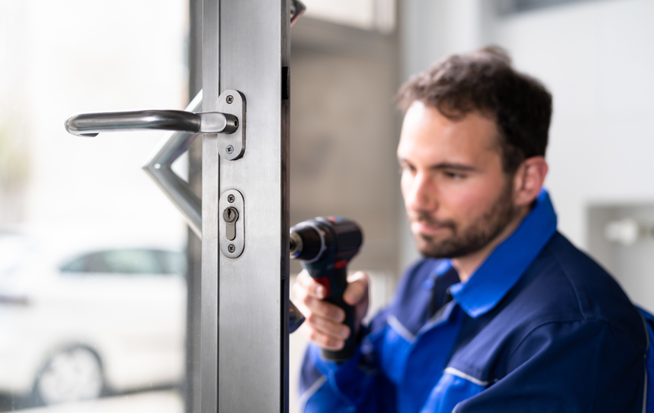 A person in blue overalls uses a power drill on a door handle near a window.