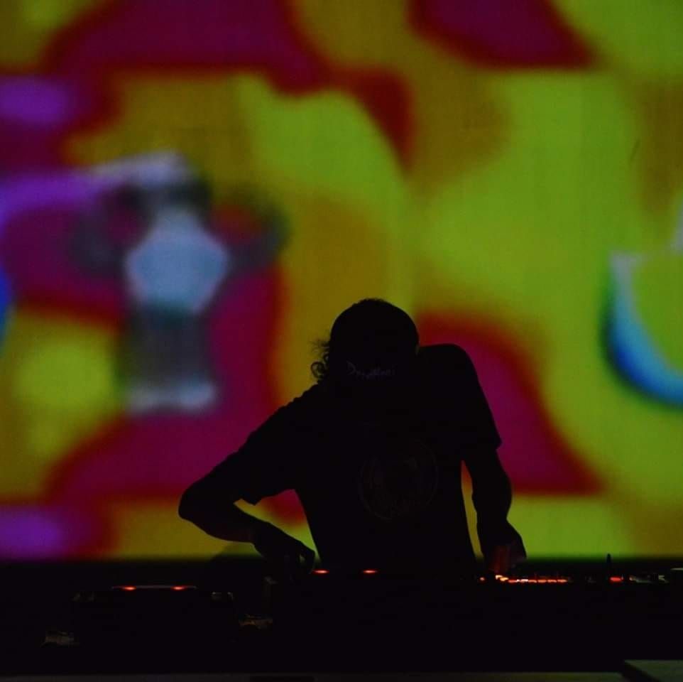 Silhouette of a DJ Curio at a mixing console with colorful abstract shapes projected behind.