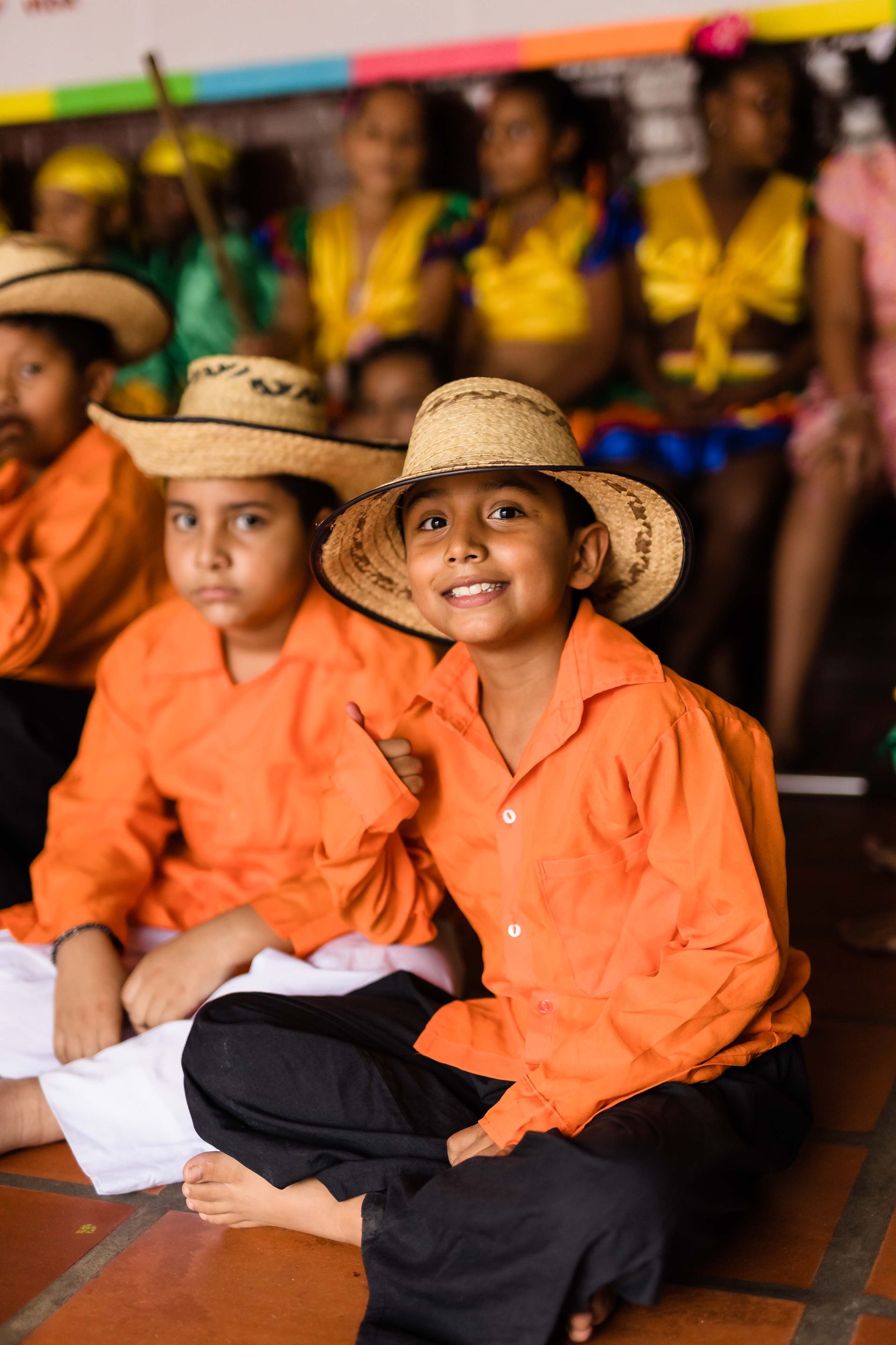A group of children wearing hats and orange shirts are sitting on the floor.