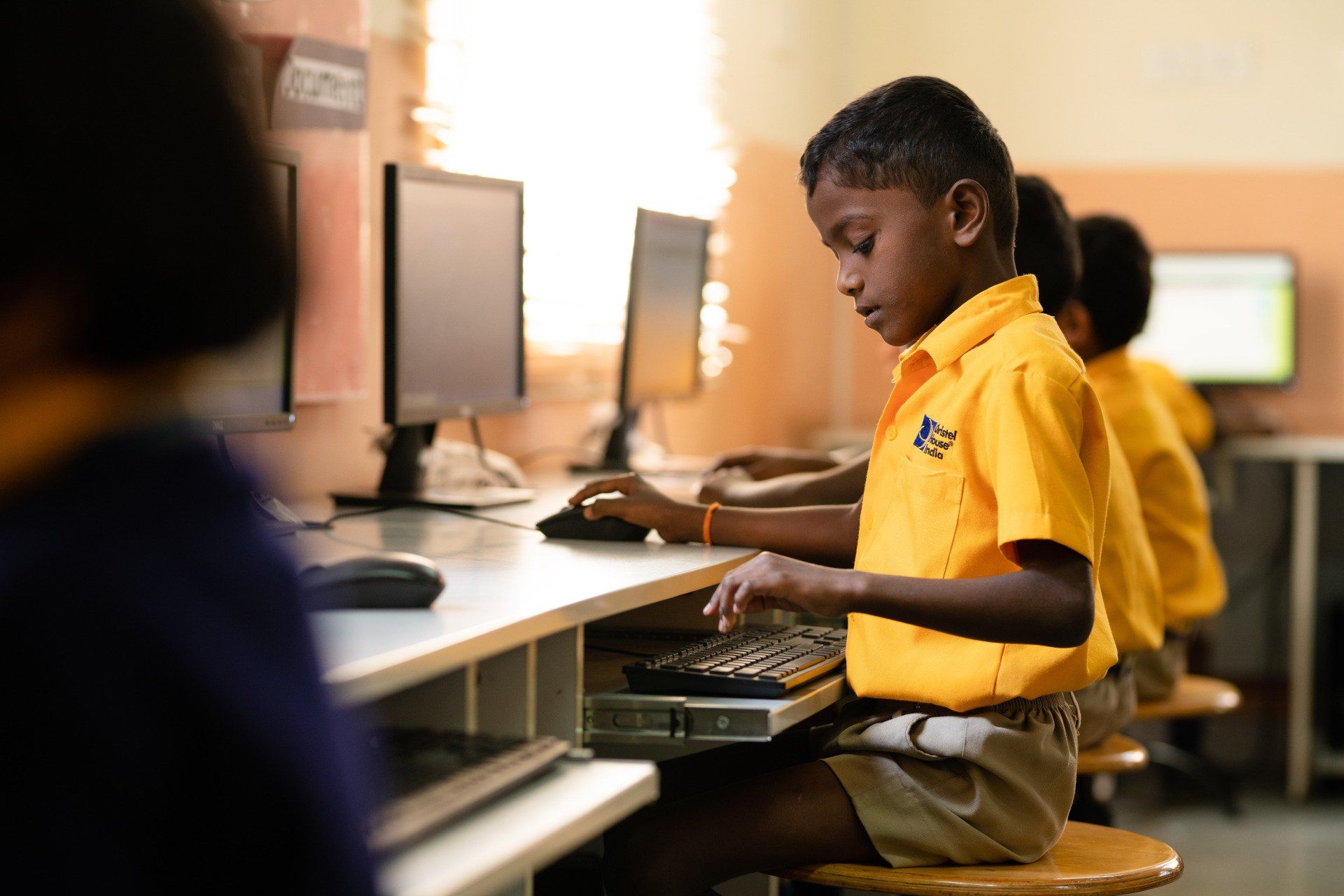 A boy in a yellow shirt is sitting at a desk in front of a computer.