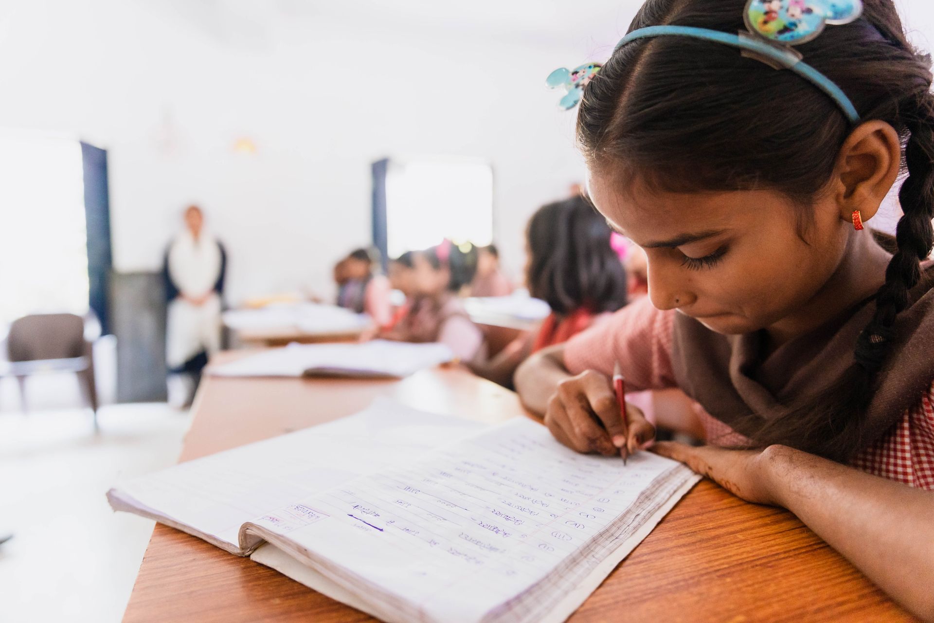 A young girl is sitting at a desk in a classroom writing in a notebook.