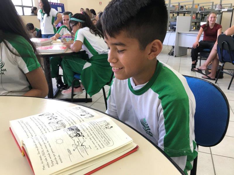 A young boy sitting in his classroom, smiling, reading a textbook. 