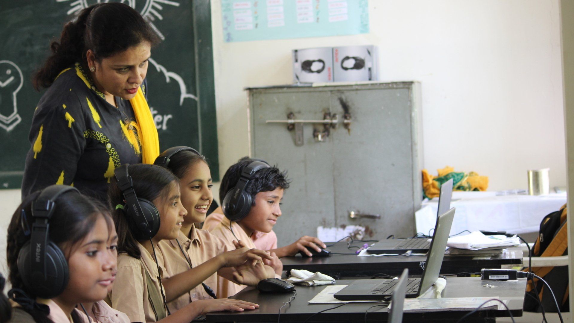 A group of children are using laptops in a classroom with a teacher standing behind them.