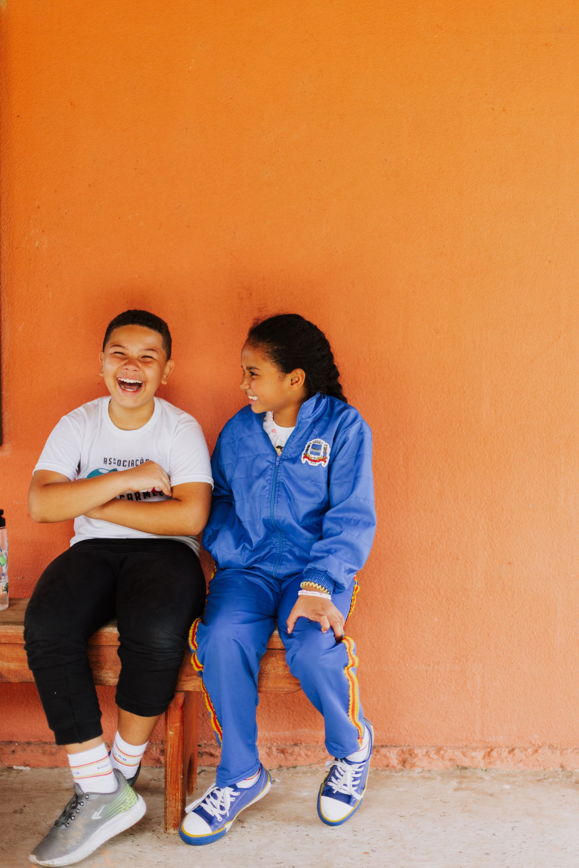 Two young girls are sitting next to each other on a bench in front of an orange wall.