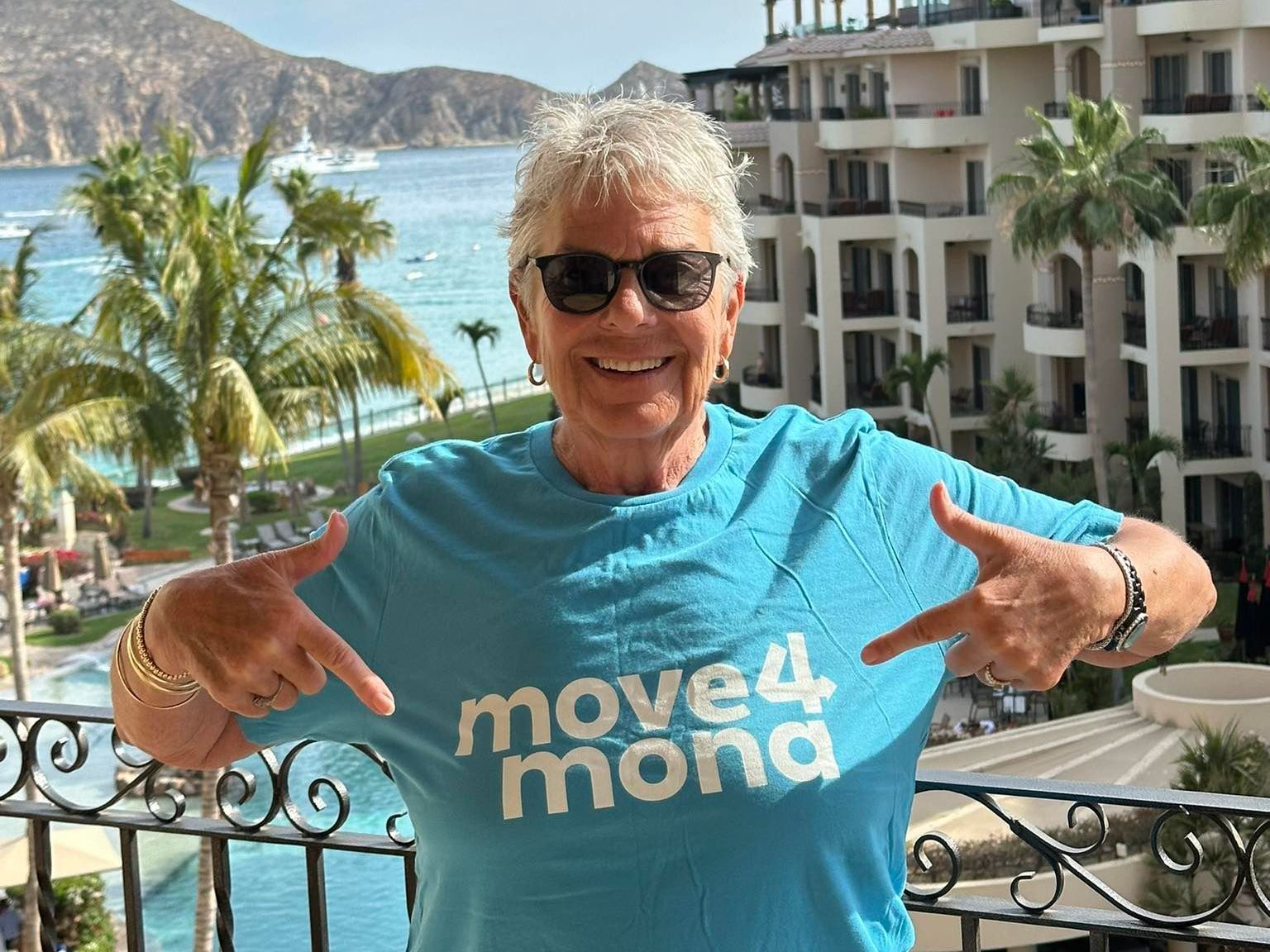 A woman wearing a blue t-shirt that says move4monda is standing on a balcony.