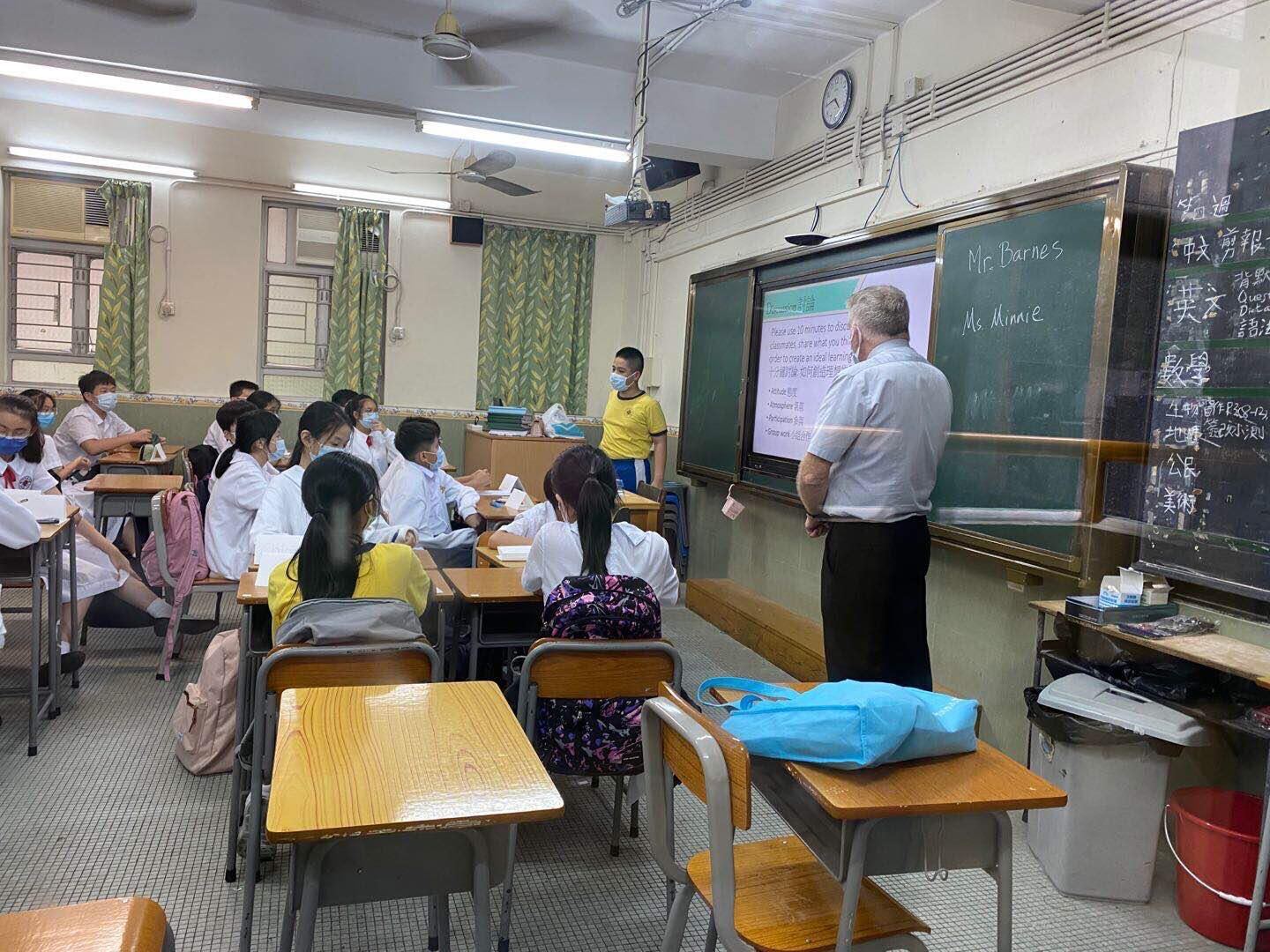 A classroom setting with students at their desks and a teacher at the front of the room using the whiteboard. 