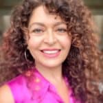 A woman with curly hair is smiling and wearing a pink shirt.