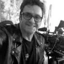 A man wearing glasses and a leather jacket is taking a selfie in front of a camera.