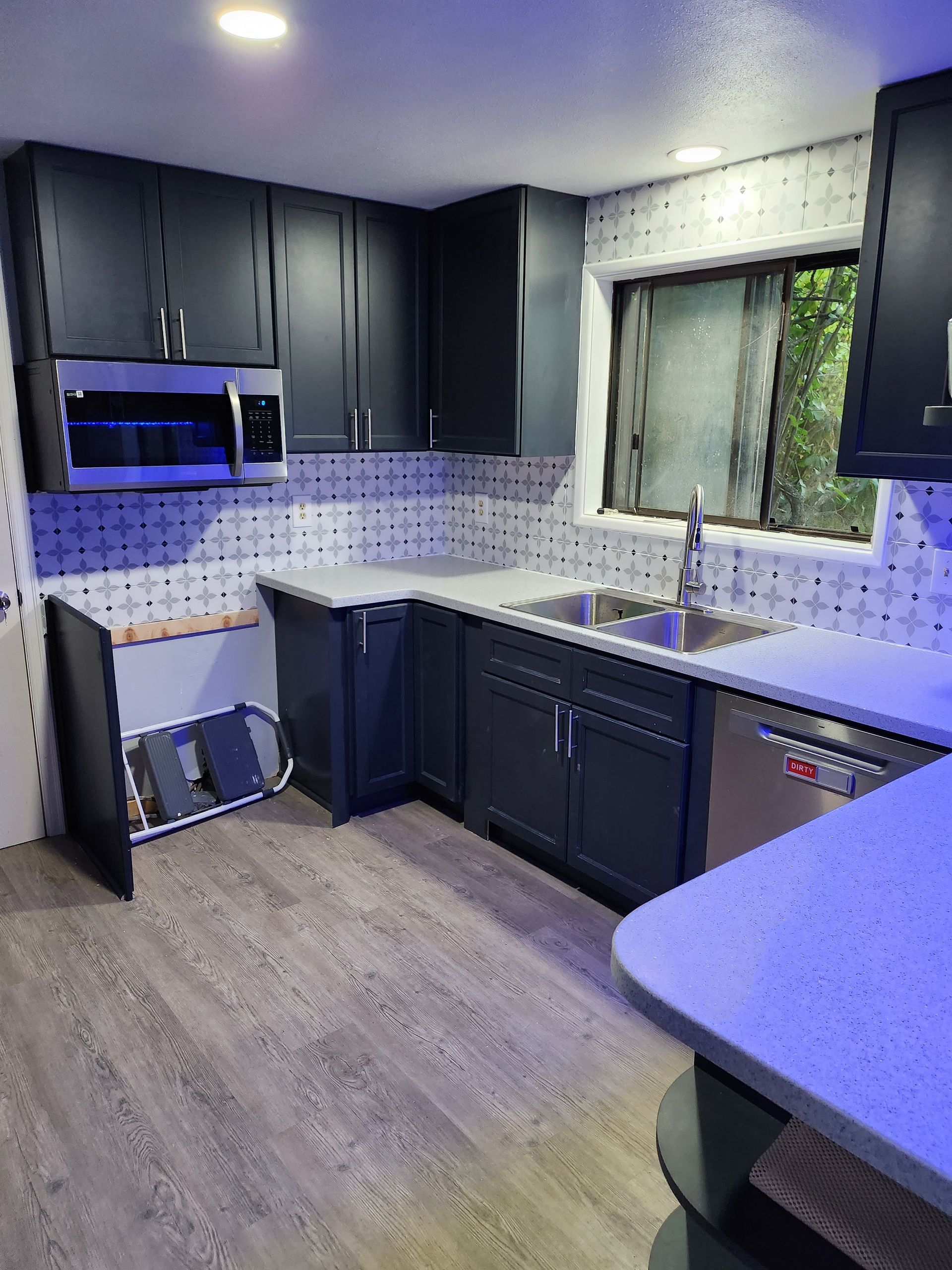 A kitchen with black cabinets , stainless steel appliances , a sink , and a window.