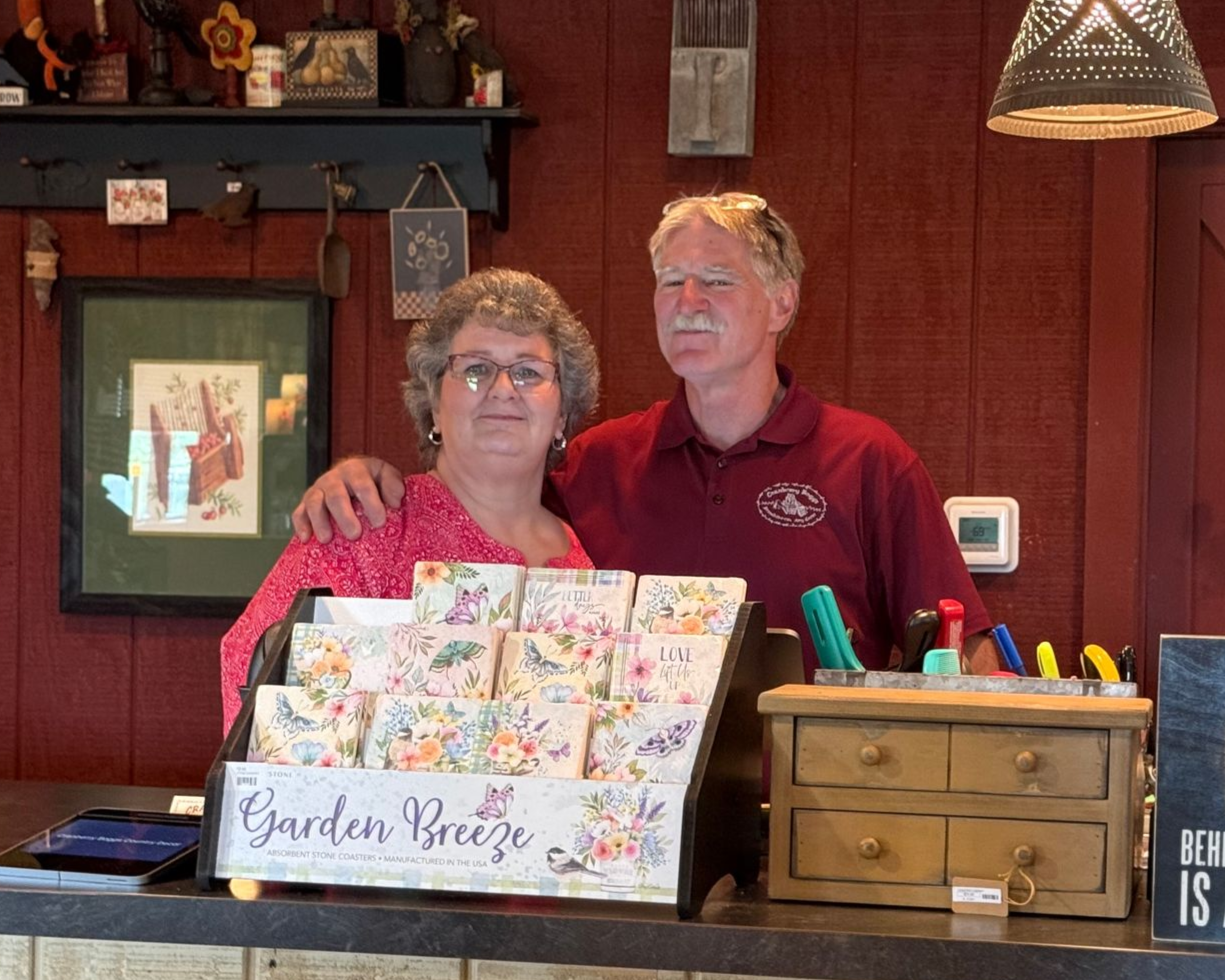 Two people behind a counter with a floral card display and a wooden cash box in a red-walled shop