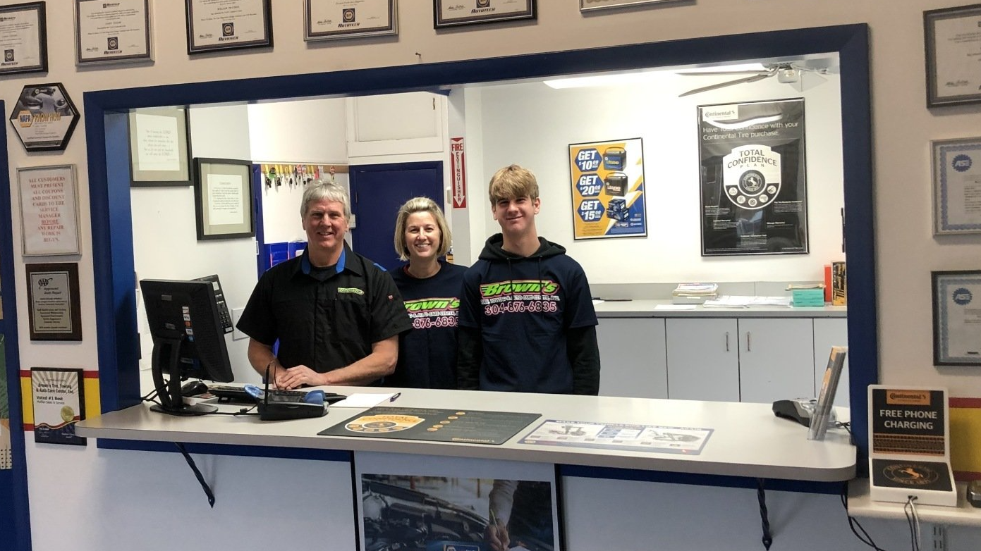 Three people at a business counter: a man, a woman, and a teen. They are smiling. The space has certificates and posters.