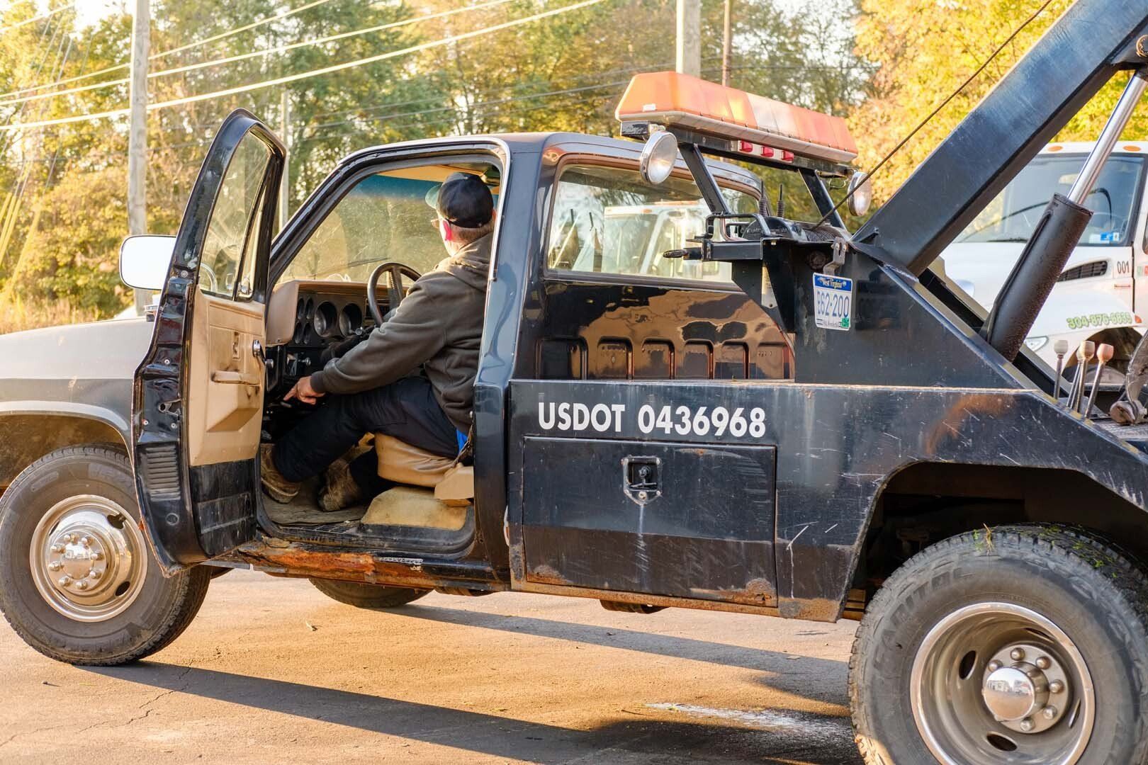 Tow truck driver sits in black tow truck with open door, parked outdoors.