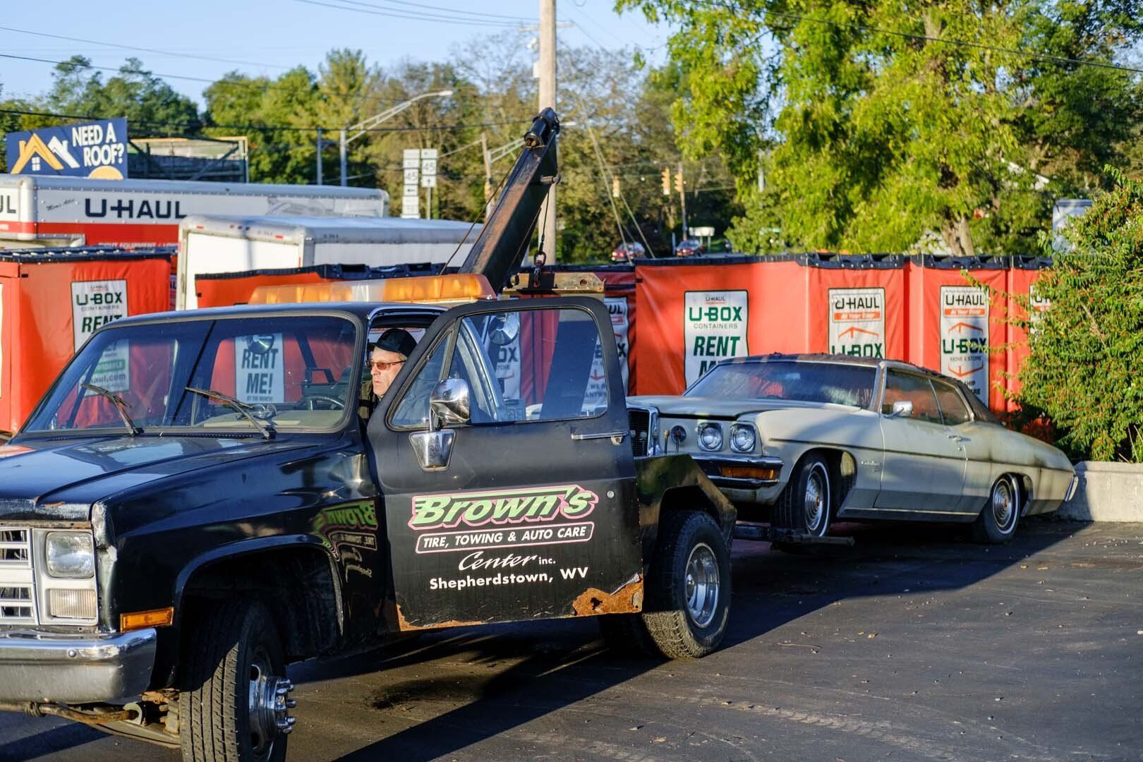 Tow truck towing a light-colored car. Truck is black with a driver. U-Haul storage units in the background.