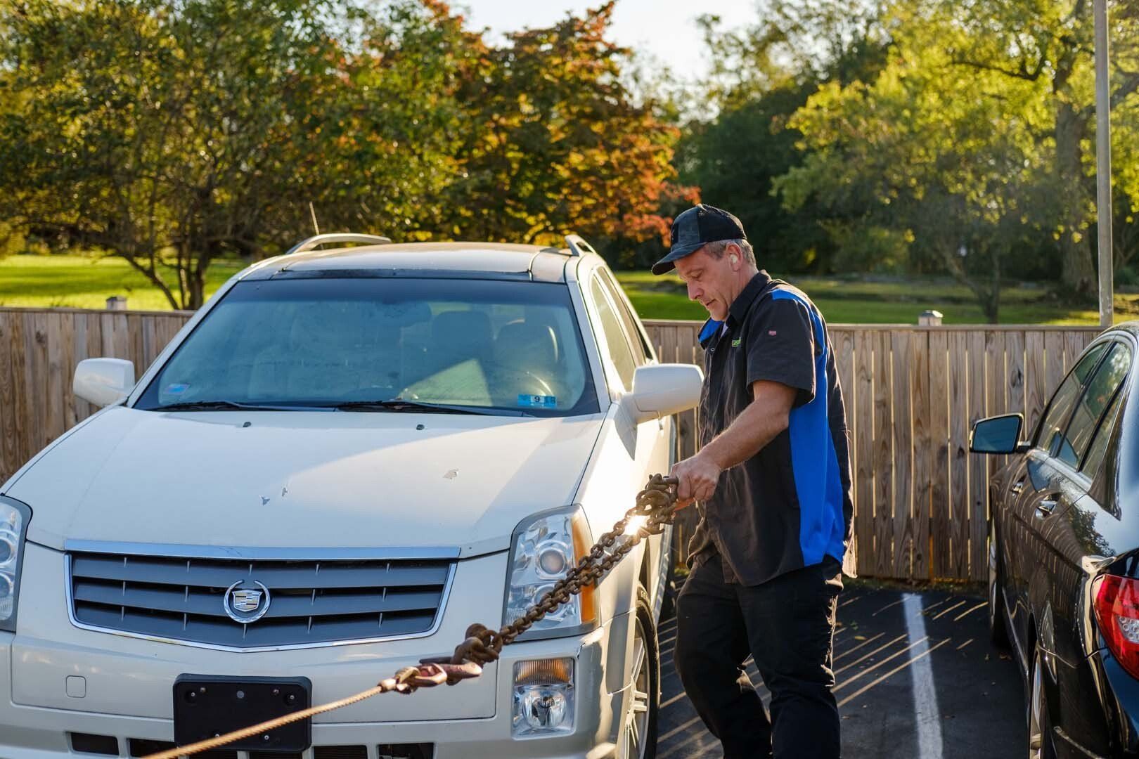 Tow truck operator connecting a tow chain to a white Cadillac SUV in a parking lot.