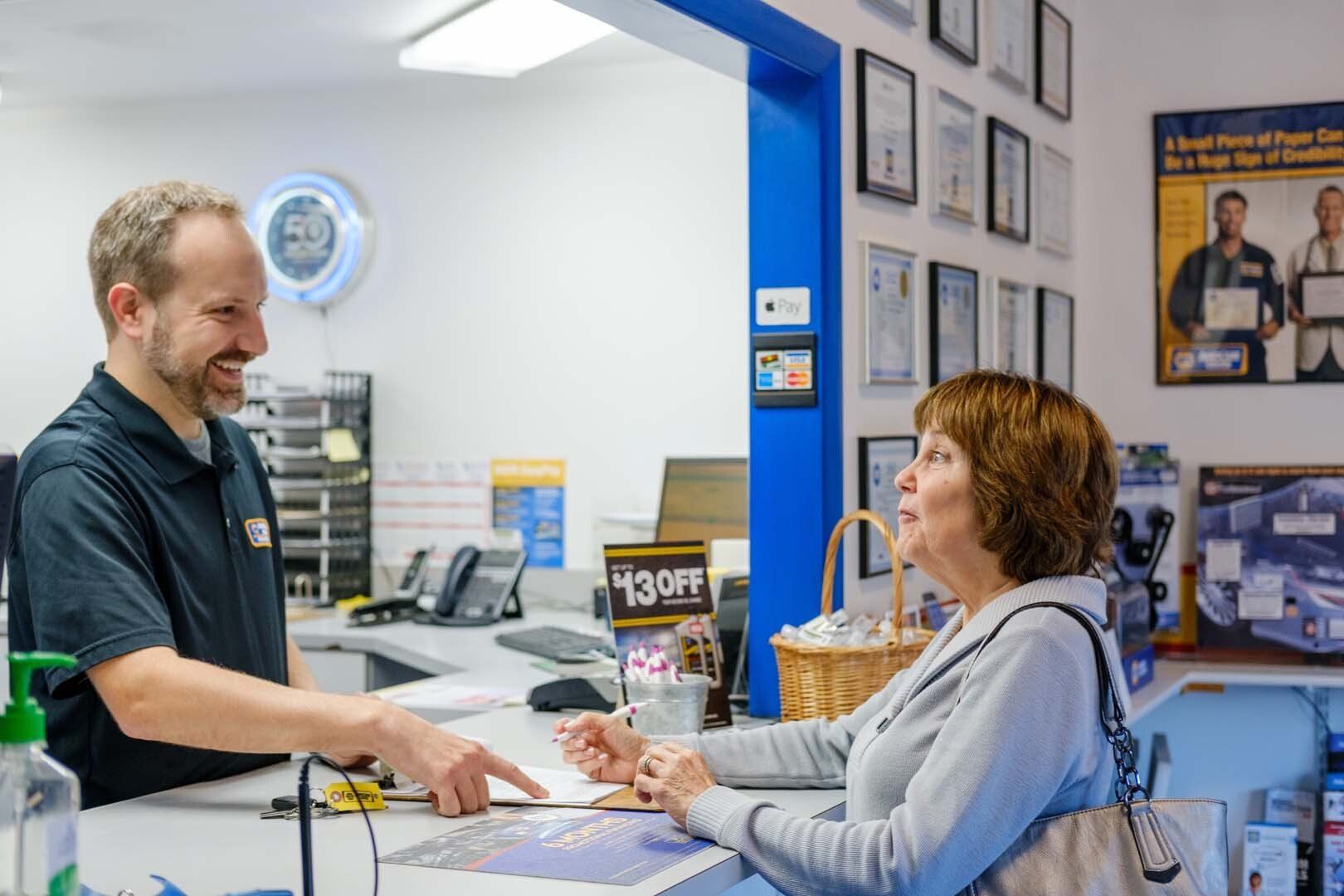 Man at car repair shop counter assisting a customer, pointing at paperwork.