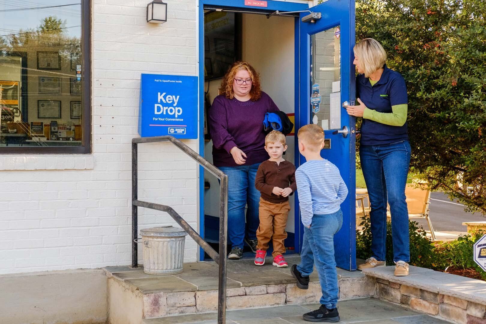 Woman opens blue door for two children, Key Drop sign beside it. Another woman looks on.