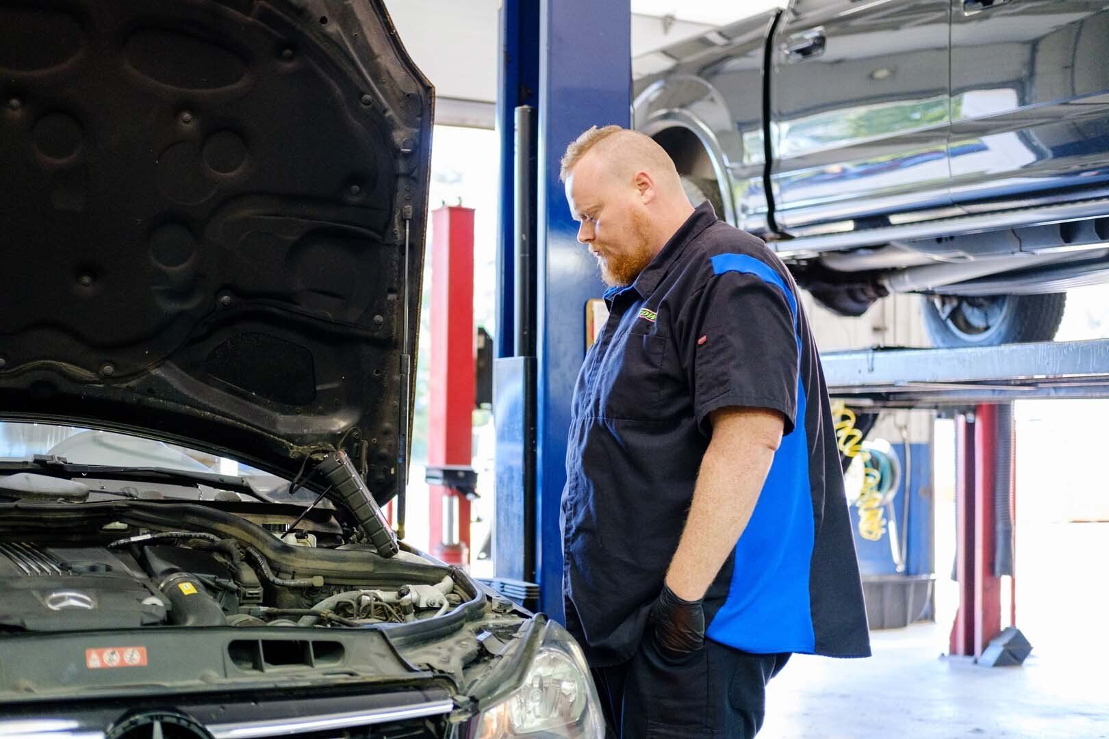 Mechanic examining a car engine with the hood open in a garage. Wearing a blue and black uniform.