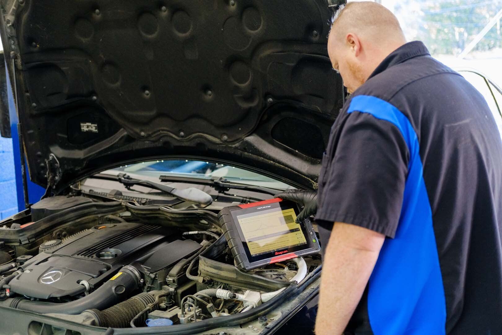 Mechanic using a diagnostic tool to examine a car engine with the hood open in a garage.
