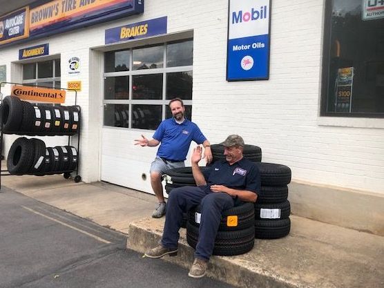 Two men at a tire shop. One sits on tires, waving; the other stands, gesturing. Exterior setting.