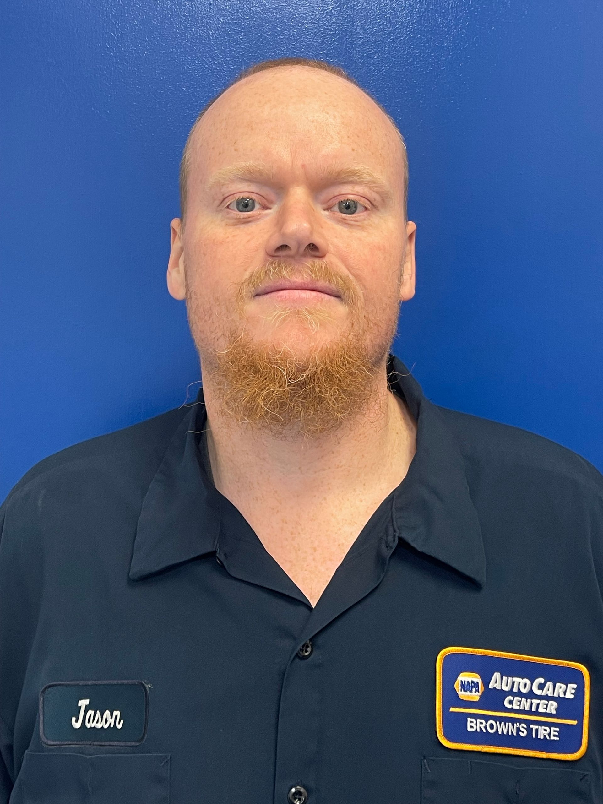 Man with red hair and beard smiles, wearing a black shirt with blue trim, against a blue background.