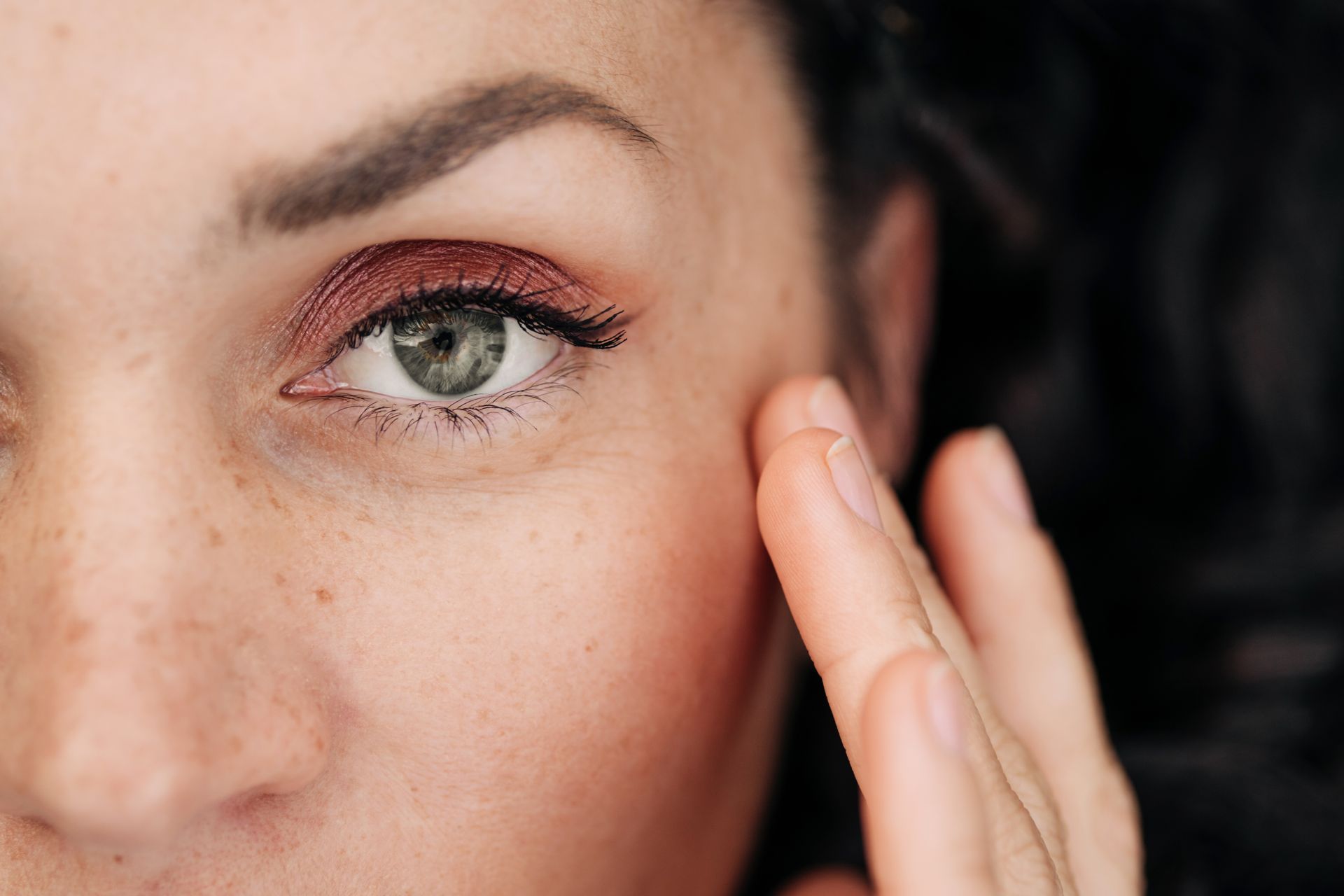 Primo piano di una donna con lentiggini e ombretto rosso, che si tocca il viso vicino all'occhio.