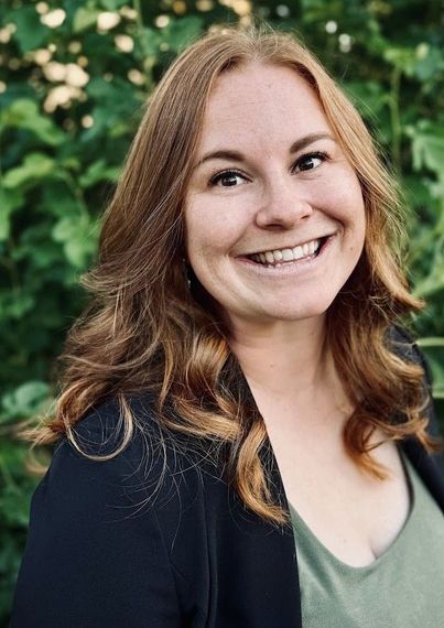 Smiling woman with long wavy hair in a dark jacket and green top outdoors with blurred greenery behind her