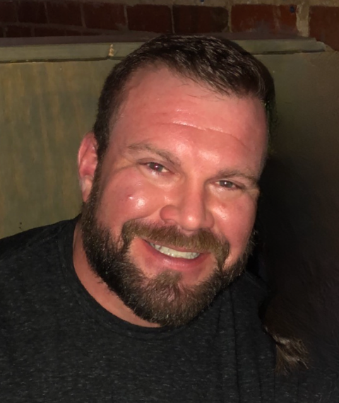 Smiling man with a dark beard and short hair, wearing a dark shirt, indoors against a brick wall.