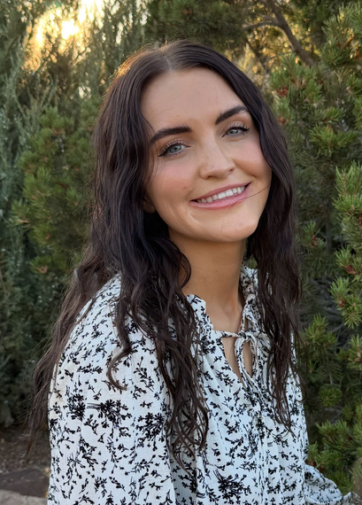 Smiling woman with long dark hair in a white patterned blouse, standing outdoors among trees.