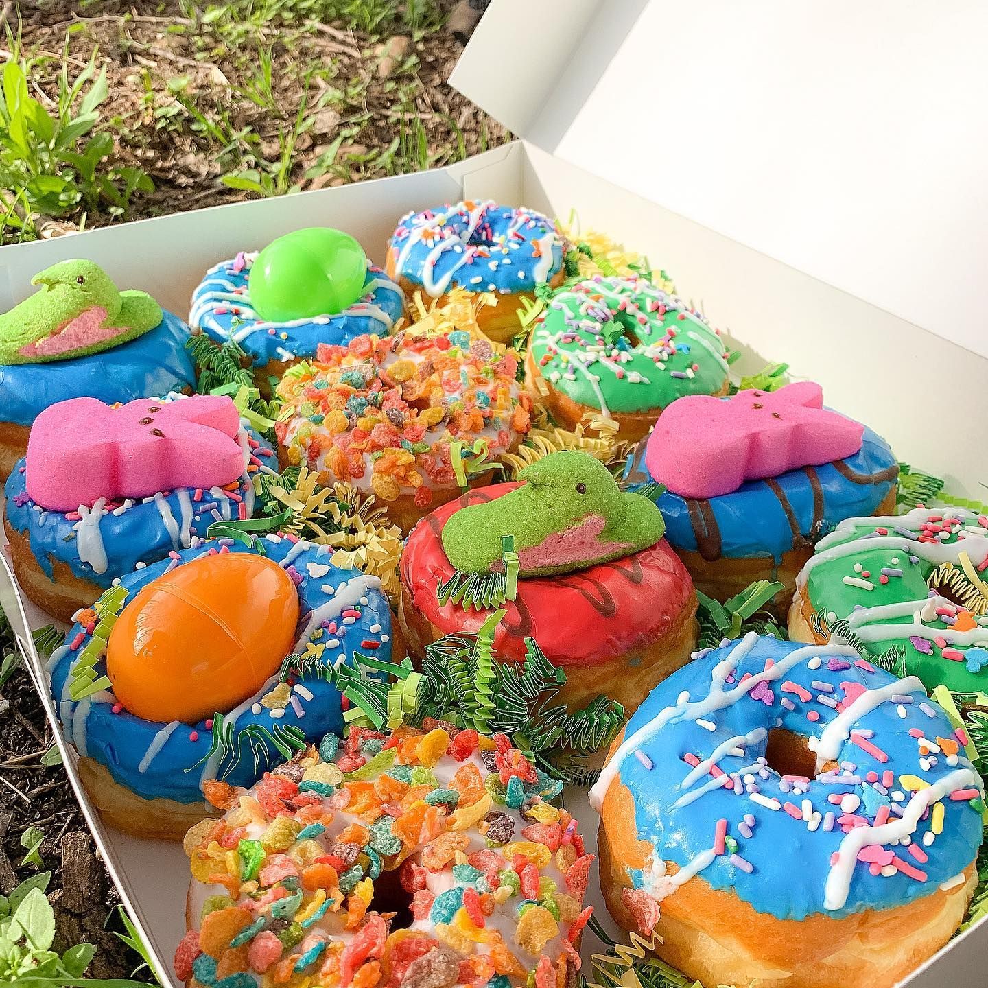 Box of colorful donuts with frosting and toppings; outdoors.