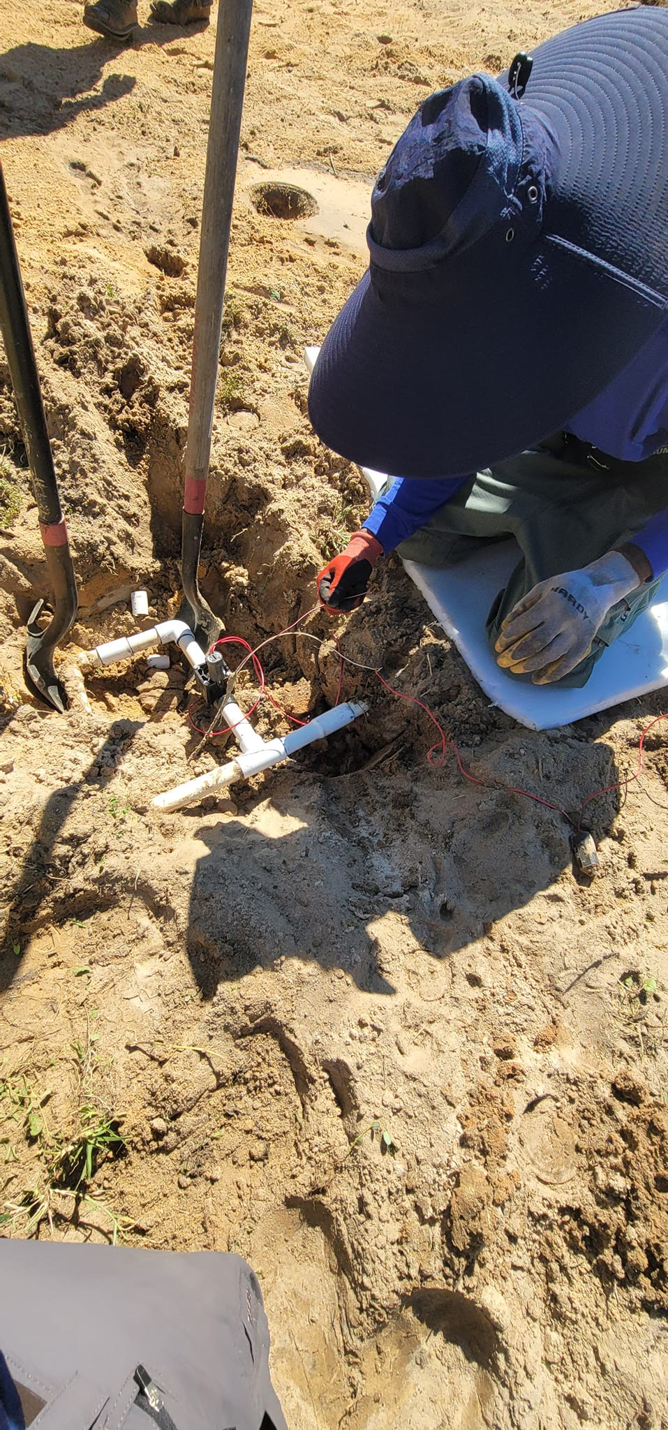 Person working on PVC pipes in a dirt trench, wearing a blue cap and gloves.
