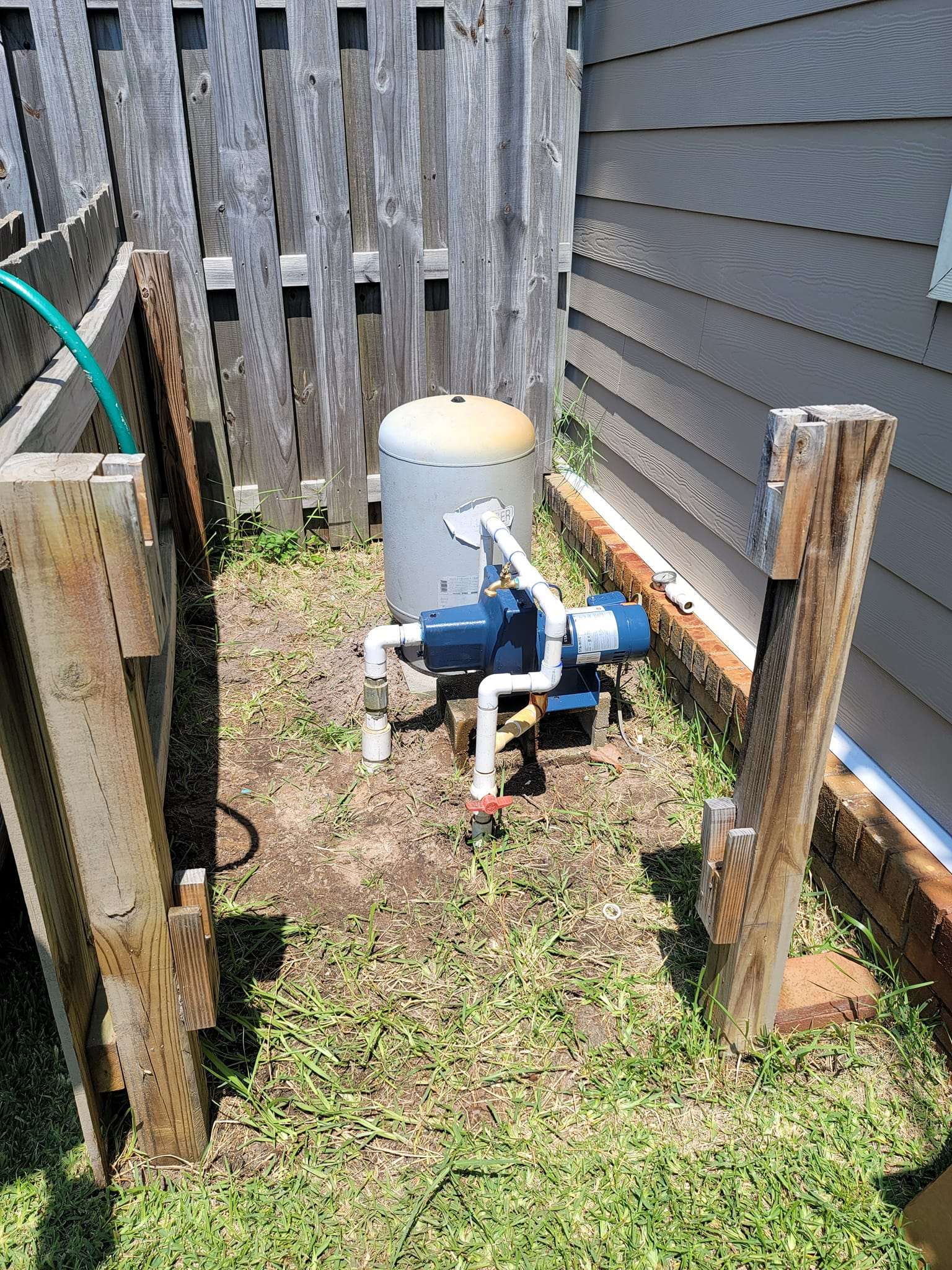Water well components: tank, pump, and piping, nestled between wooden fencing and siding, on grass.