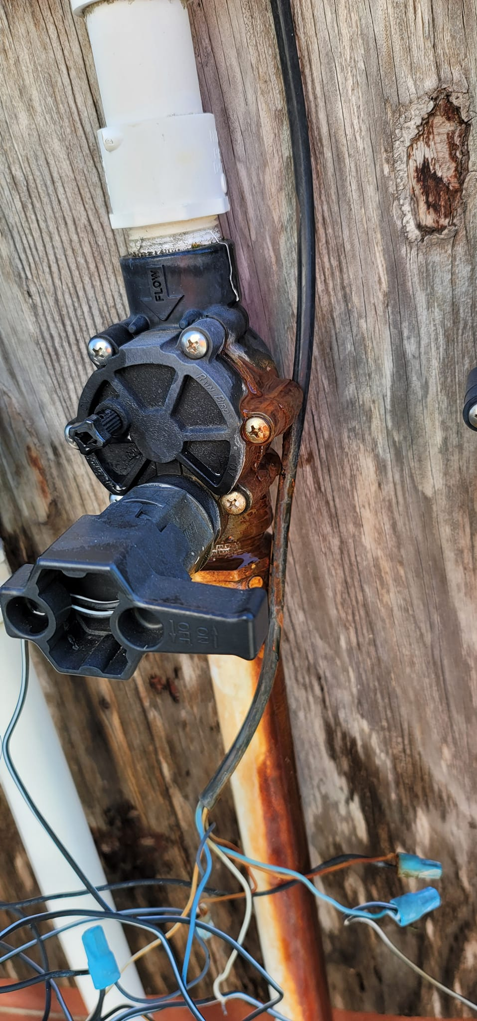 Close-up of a black irrigation valve attached to wood, with wires and white PVC pipe visible.