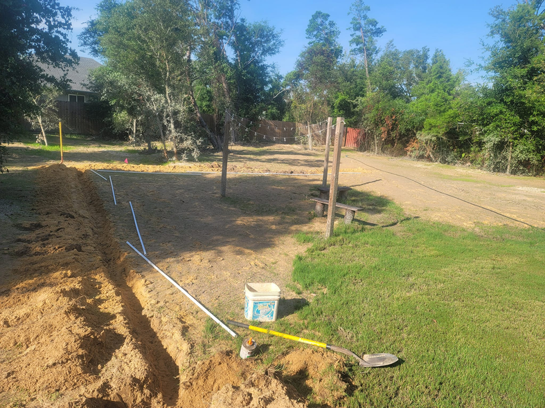 Trench dug in yard, white pipe and tools beside it. Wooden posts and trees in the background. Sunny day.